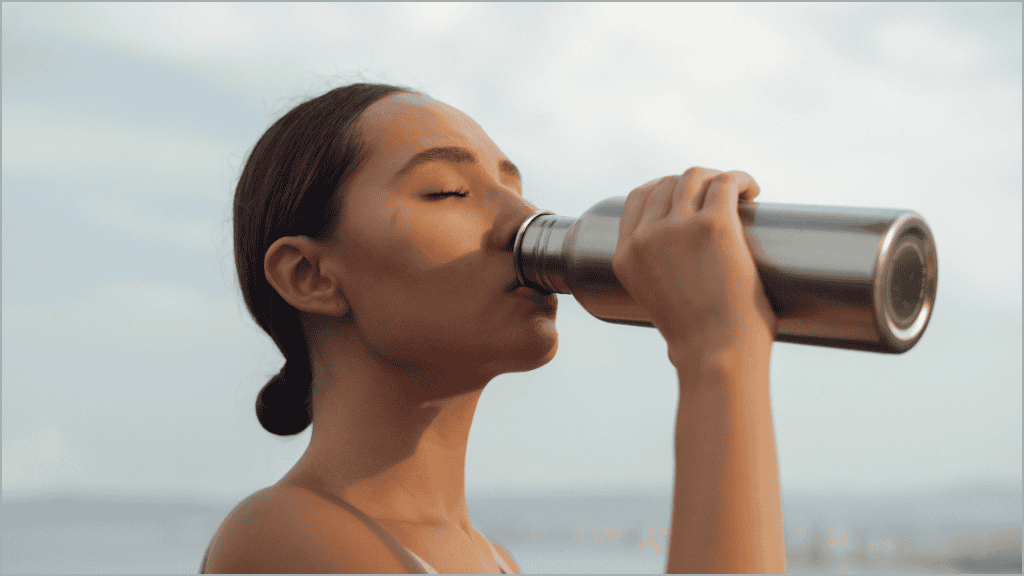 A woman in a serene outdoor setting drinks deeply from a stainless steel water bottle, her eyes closed in a moment of refreshment.