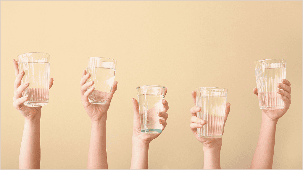 Multiple hands holding up clear glasses of water against a soft yellow background, symbolizing community and shared commitment to hydration.