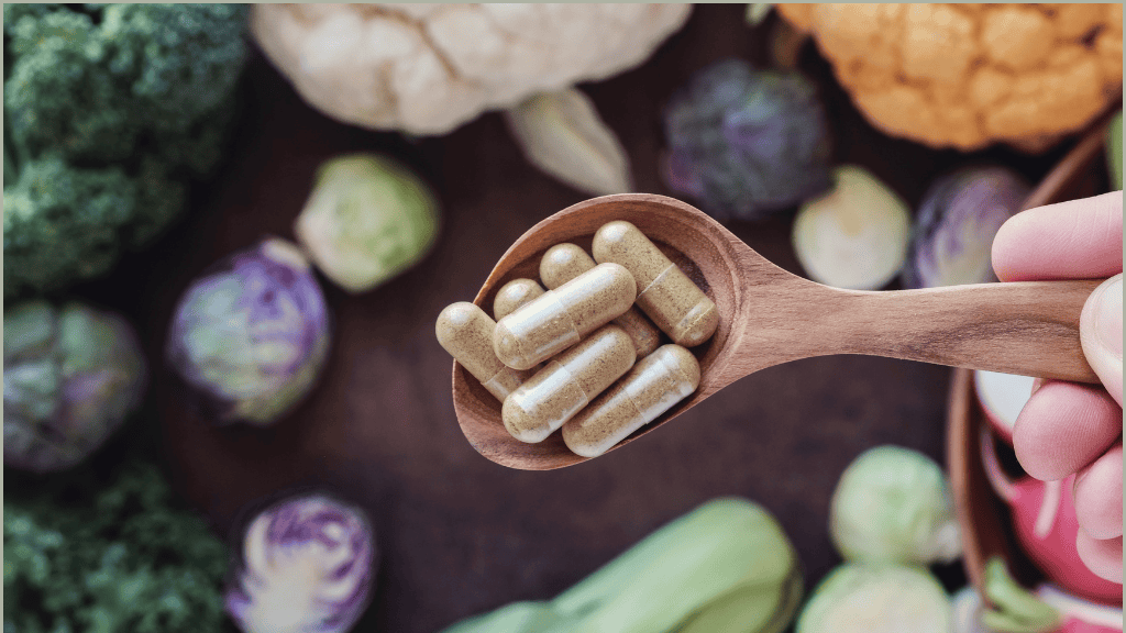 Close-up of a wooden spoon held over a variety of fresh vegetables, containing capsules filled with natural supplements, illustrating the connection between dietary choices and supplemental nutrition.