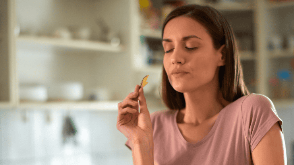 A woman enjoying a moment of mindful eating, savoring a chip with her eyes closed, in a home kitchen setting.