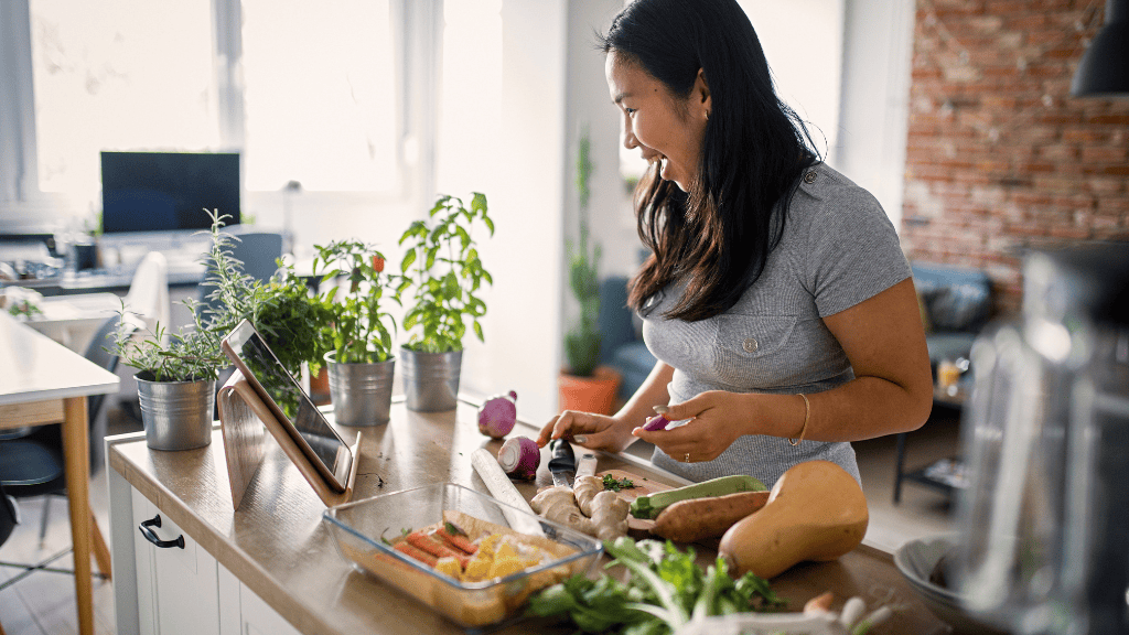 A woman joyfully prepares vegetables in a kitchen filled with natural light, with fresh herbs in pots and a digital recipe guide on her tablet.