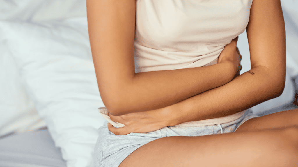 A woman sitting on a bed, holding her abdomen gently.