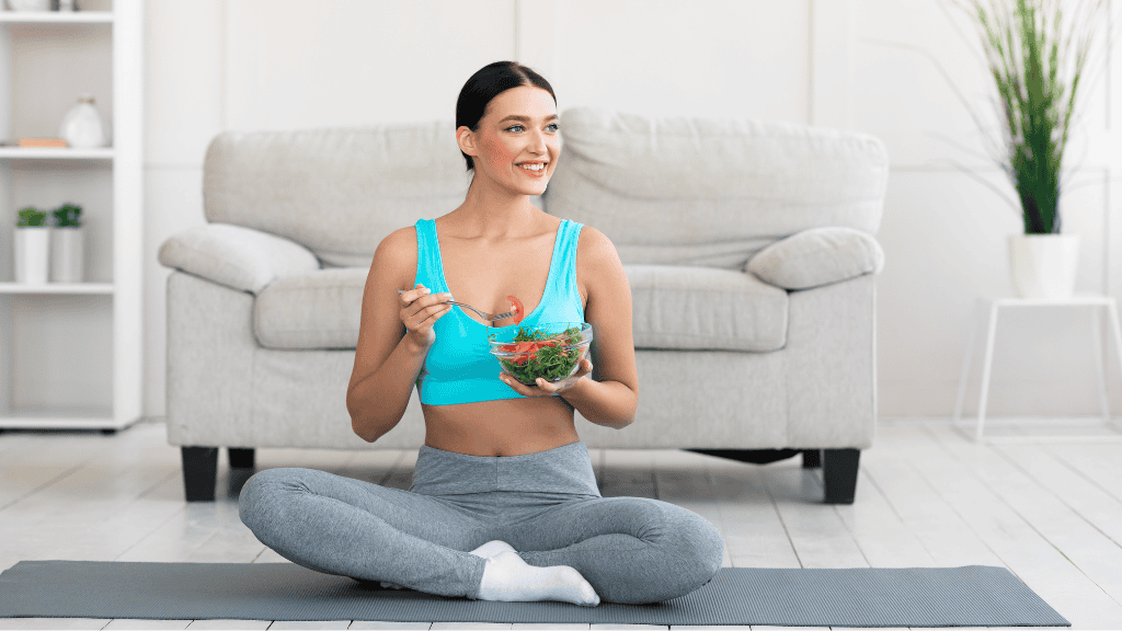 A young woman in workout gear sits cross-legged on a yoga mat in her living room, eating a fresh salad and smiling.