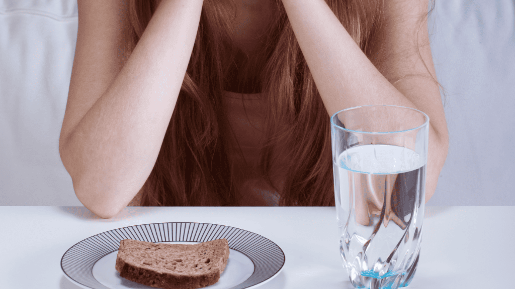 A young woman sits at a table, head resting on her arms, facing a simple meal of bread and water, reflecting the struggle with restrictive eating.