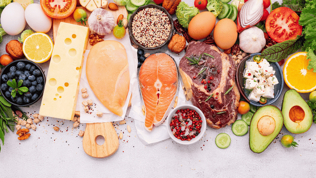 A colorful array of various whole foods including fruits, vegetables, nuts, cheeses, and meats spread out on a white surface.