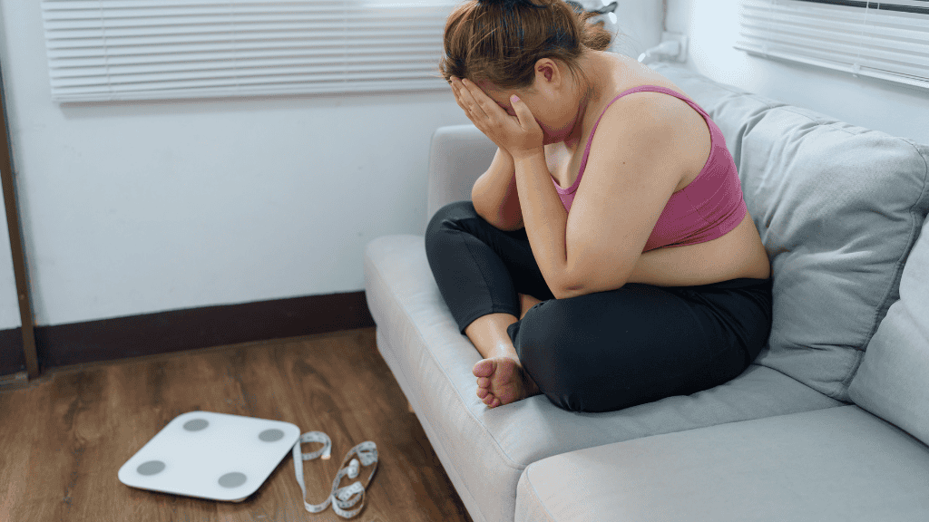 A woman in workout attire sits curled up on a couch, distressed, with a scale and measuring tape nearby, reflecting the emotional impact of dieting.