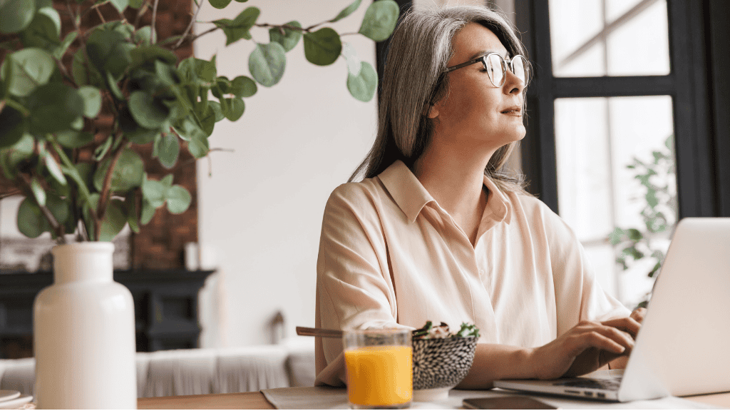 A mature woman in a light pink blouse sits at a dining table, looking thoughtful as she works on her laptop surrounded by lush indoor plants.