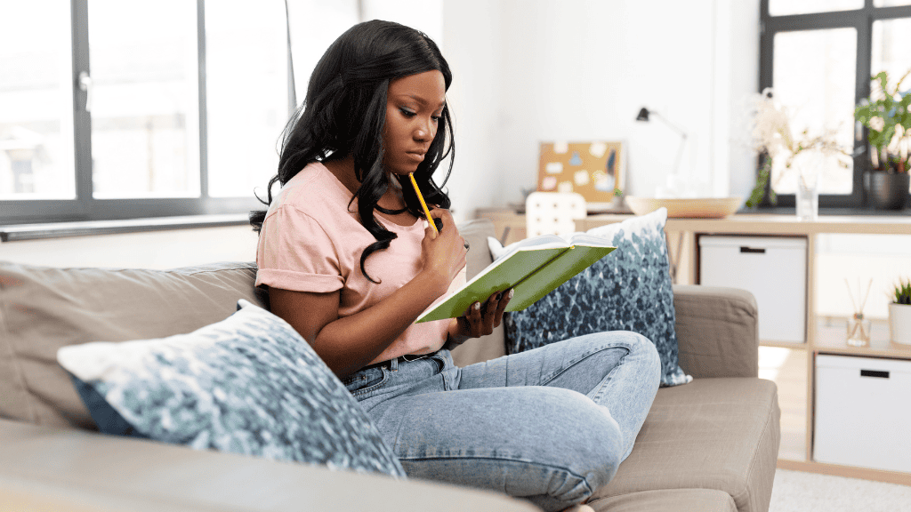 A focused young woman sits on a sofa, deeply engaged in writing in her journal, pencil in hand.