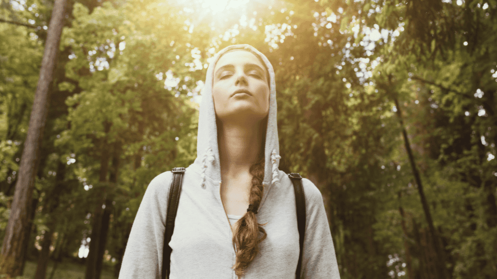 A serene woman with a hoodie, standing in a forest, looks upward with a peaceful expression, bathed in sunlight filtering through the trees.