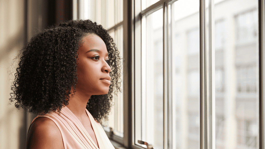 A young woman with curly hair gazes thoughtfully out a window in a well-lit room.