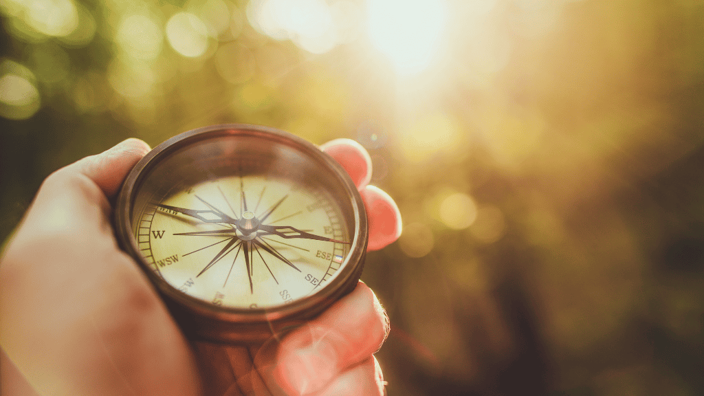 A hand holding a compass in a sunlit forest, focusing on the needle pointing east.