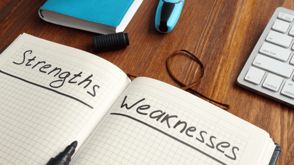 An open notebook with the words "Strengths" and "Weaknesses" written on opposite pages, alongside a keyboard and other work tools on a wooden desk.