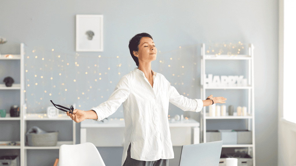 A woman in a white shirt stands in a tranquil home office, eyes closed and arms outstretched, embodying a moment of peaceful reflection.