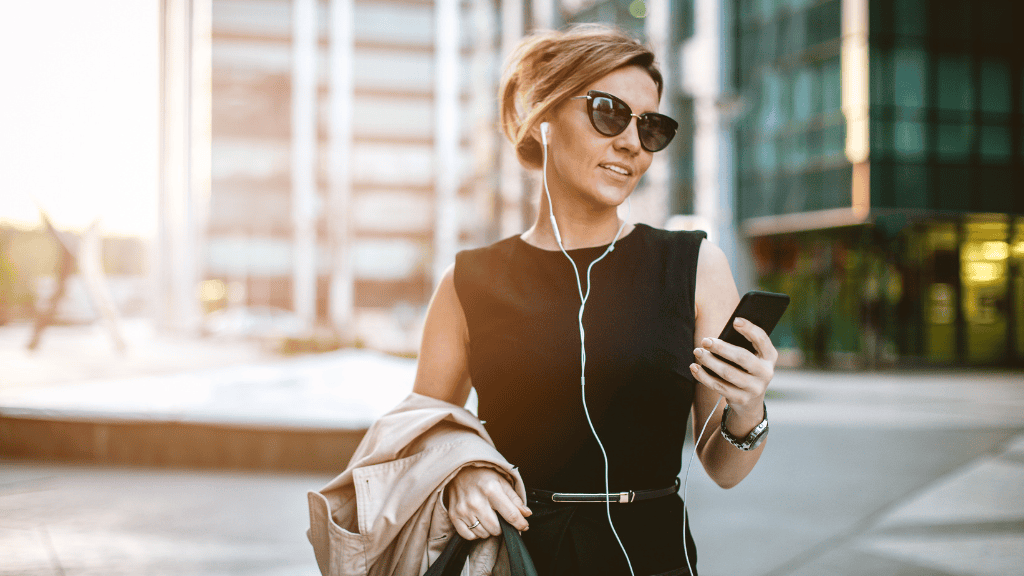 A busy woman in her mid-thirties wearing sunglasses and a black sleeveless top walks confidently through a sunny urban area, listening to music on her phone.