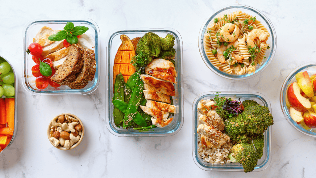 An assortment of healthy meals in glass containers arranged on a marble countertop, featuring fruits, vegetables, nuts, grains, and proteins.