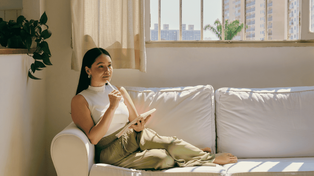 A thoughtful woman sitting on a couch by a sunny window, holding a notebook and a pen, deep in planning.