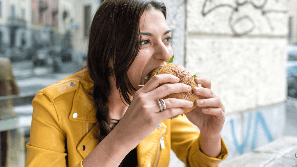 A woman in a vibrant yellow jacket eating a  sandwich at a street-side café.