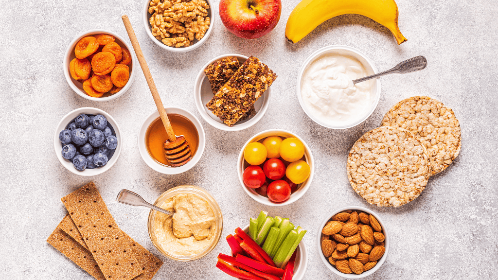 An assortment of healthy snacks neatly arranged on a gray surface, including fruits, nuts, yogurt, and whole grains.