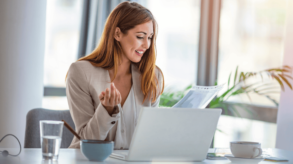 A happy businesswoman reviewing documents while working on her laptop at a bright and airy office, a bowl of healthy snacks nearby.