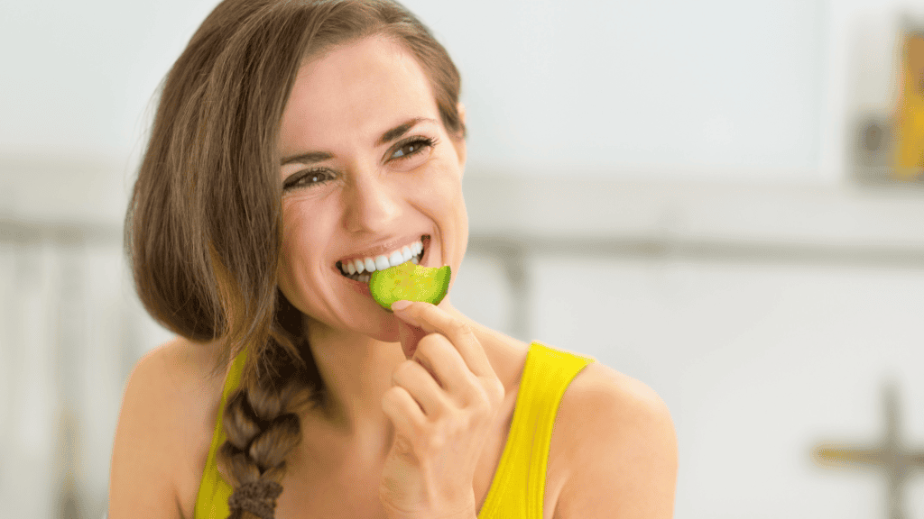 A joyful woman in a yellow tank top enjoying a slice of cucumber, embodying a healthy lifestyle.