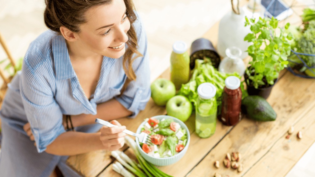 A smiling woman enjoys a fresh salad amidst a spread of healthy foods and drinks on a wooden table.