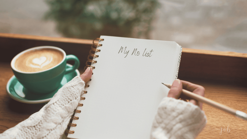 Practicing self-care, a women in a cozy sweater writes "My No List" in a notebook beside a cup of coffee with a heart-shaped design on a wooden table.