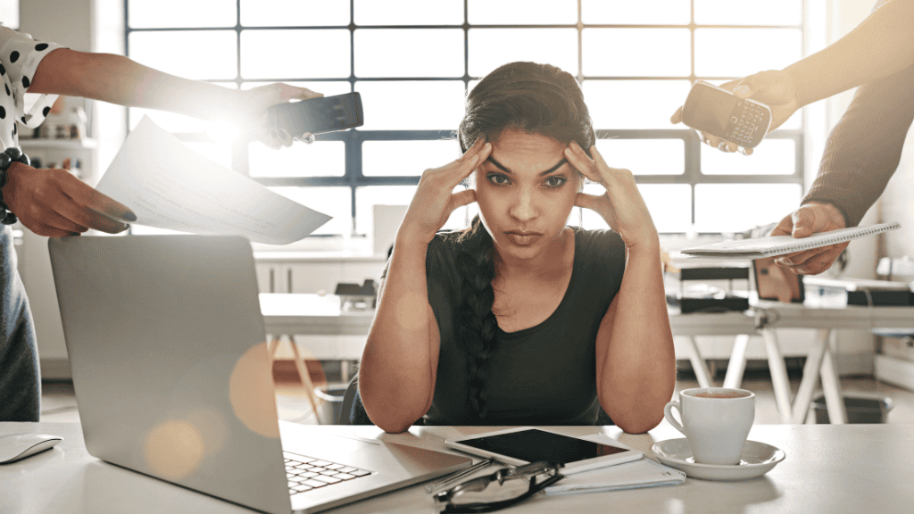 A stressed woman at her desk surrounded by coworkers offering her documents, a phone, and a laptop.