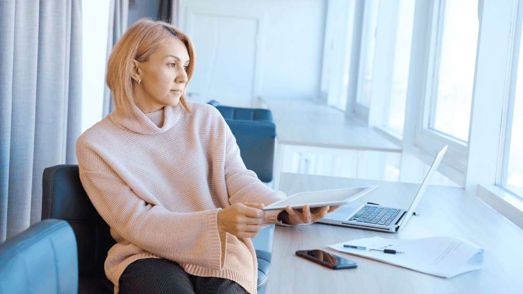 A thoughtful woman in a pink sweater uses a tablet while sitting in a bright, modern office.