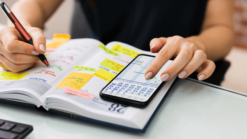 Close-up of a woman's hands writing in a planner filled with sticky notes and using a smartphone, emphasizing detailed scheduling.