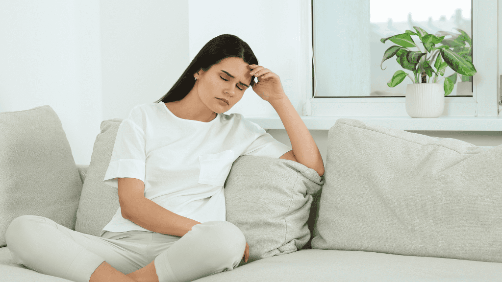 A young woman sitting on a couch, holding her forehead in discomfort, with a potted plant in the background.