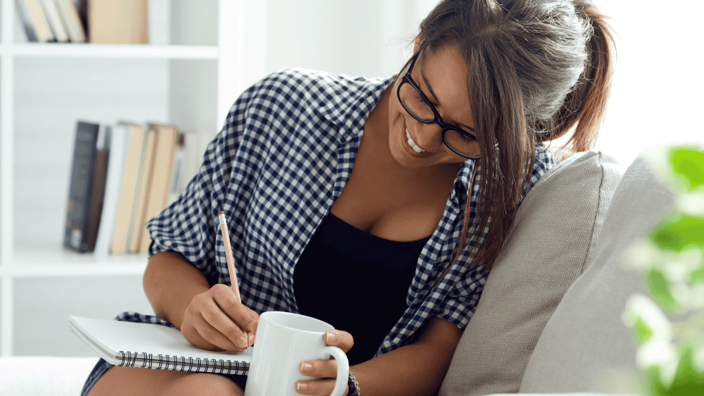 A smiling woman in glasses writing in a notebook at home, looking content and focused.