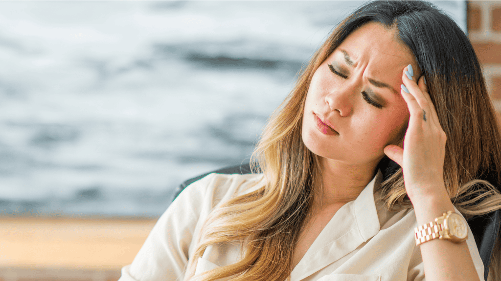 A woman sitting in an office, looking stressed with one hand on her forehead, embodying the need for self-care and well-being.