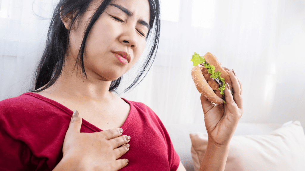 Woman experiencing discomfort while holding a hamburger, illustrating the impact of poor dietary choices on physical well-being.