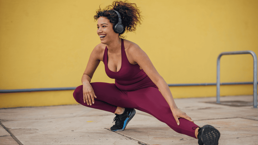 Happy woman doing a stretch exercise in workout clothes, enjoying her fitness routine outdoors against a bright yellow wall.