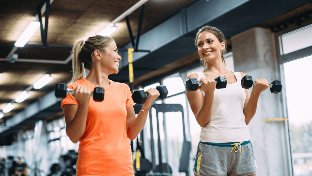 Two women happily lifting dumbbells in a gym, engaged in a conversation while exercising. Both are wearing workout clothes, smiling, and displaying positive energy.