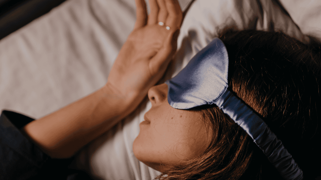 A woman wearing a sleep mask lies in bed, her hand resting on her forehead, embodying a moment of peaceful rest in a serene environment.