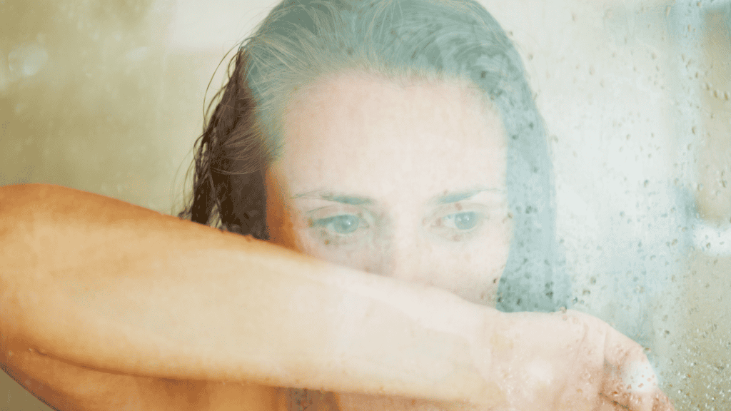 A woman behind a steamy glass shower, her arm shielding her face, embodies the weight of chronic stress.