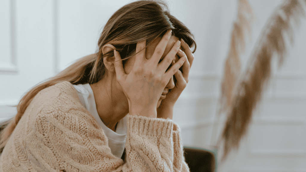 A distressed woman sits with her hands covering her face, signaling overwhelming stress or sorrow in a serene indoor setting.