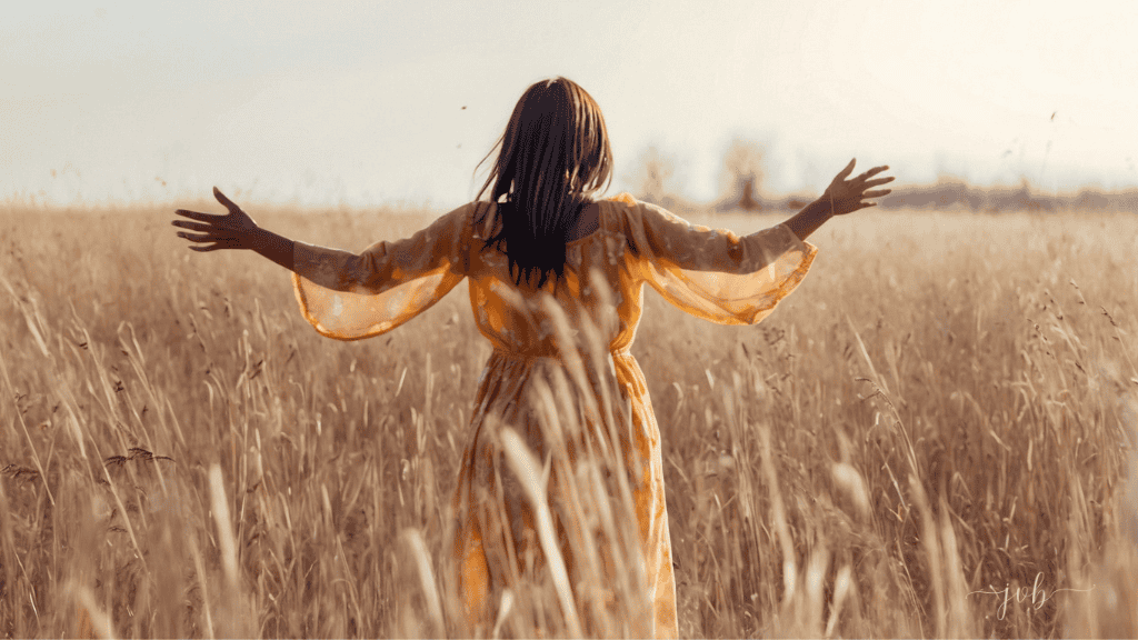 A woman in a flowing yellow dress joyfully spreads her arms wide in a golden wheat field, celebrating her connection with nature.