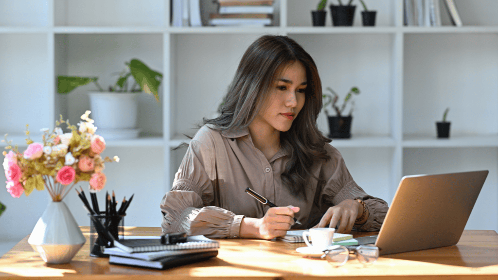 A focused woman works at her laptop in a well-organized workspace, surrounded by houseplants and a vase of flowers, illustrating a serene and productive environment.