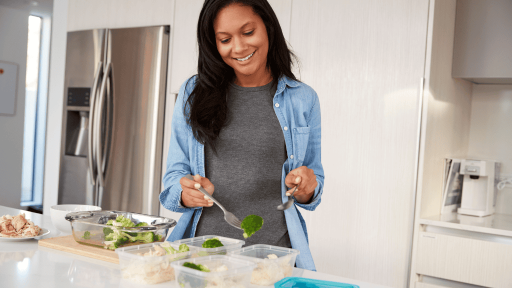 A smiling woman in a blue denim shirt preparing a healthy meal with broccoli and other ingredients in her kitchen.