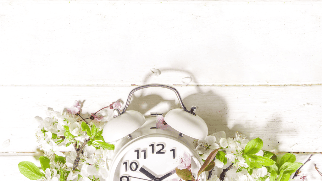 A white alarm clock surrounded by spring flowers on a white wooden surface, casting a shadow.