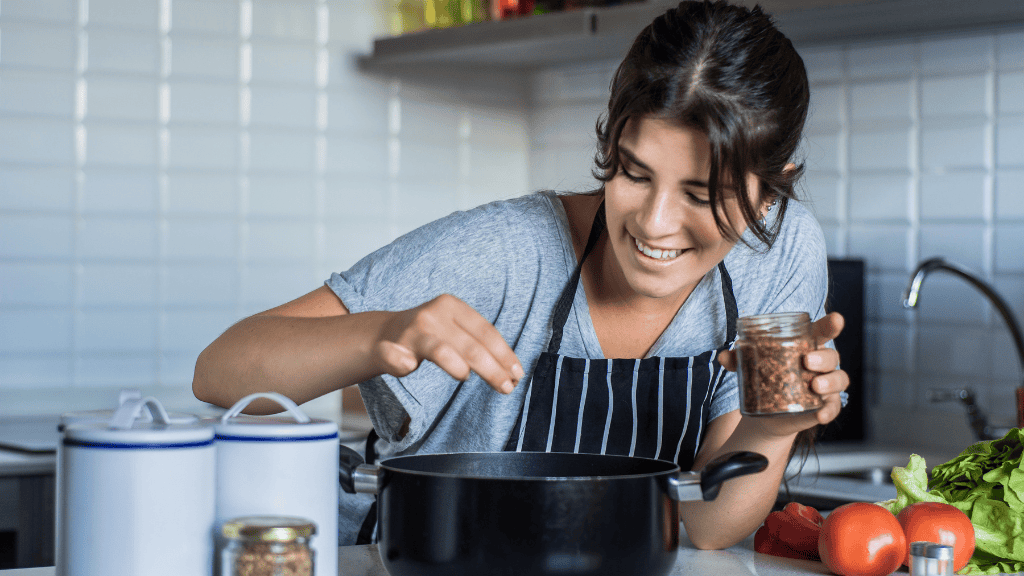 A smiling woman in a gray sweater adding ingredients to a slow cooker in her kitchen, surrounded by vegetables and jars.