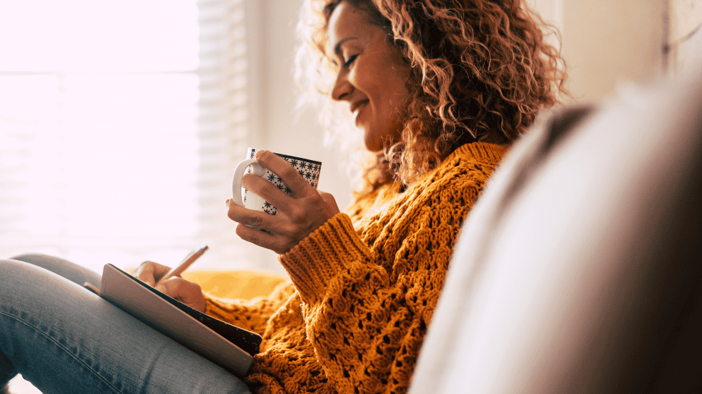 Smiling woman in a cozy sweater jotting down notes in her notebook, planning meals while sipping tea in a sunny room.