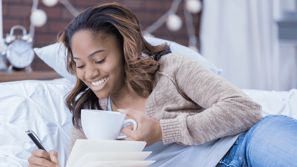 Happy woman writing in her notebook while enjoying a cup of coffee in bed, planning her meals.