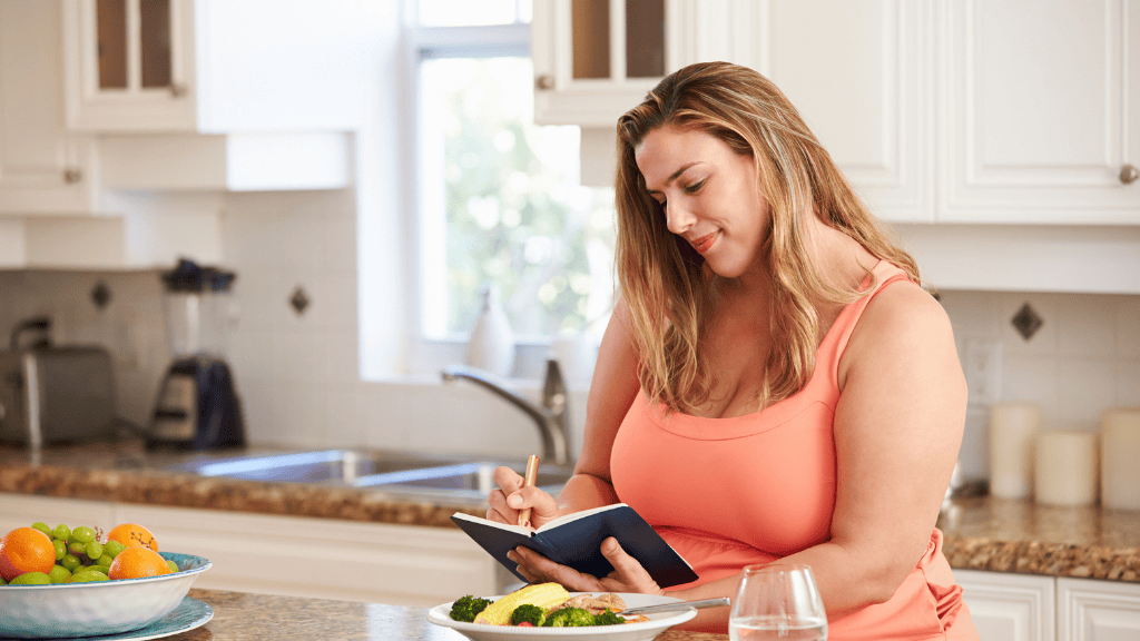 Woman in a bright kitchen, writing in a notebook while enjoying a healthy meal, embodying a relaxed meal planning routine.