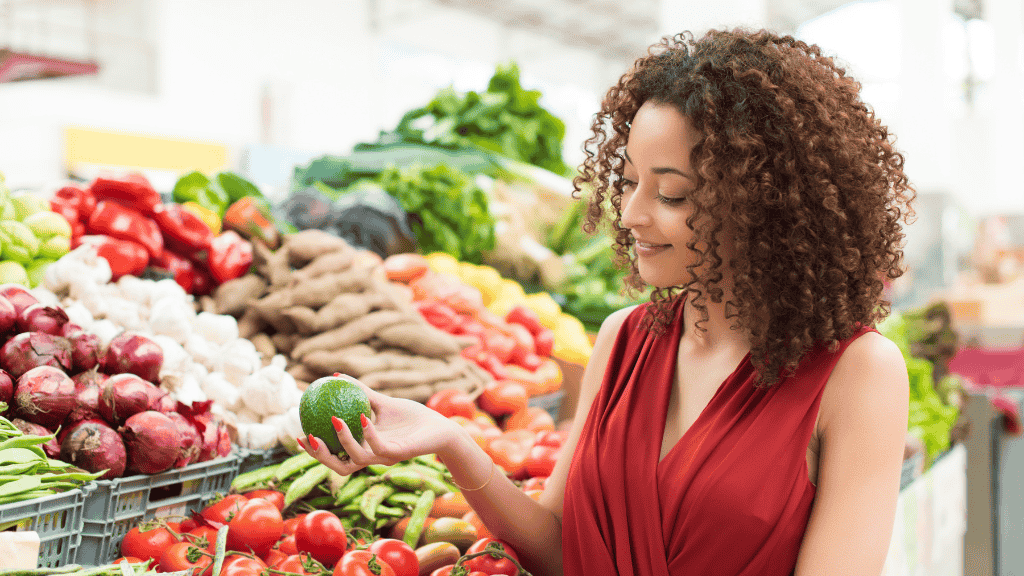 Woman carefully selecting an avocado at a bustling farmers' market, surrounded by a variety of fresh vegetables.