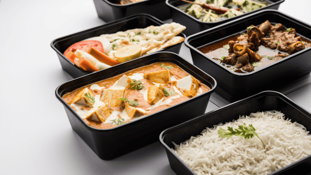 Assorted meals in black takeaway containers including butter paneer, mutton curry, and steamed rice, served with fresh sliced vegetables and naan bread on a white background.