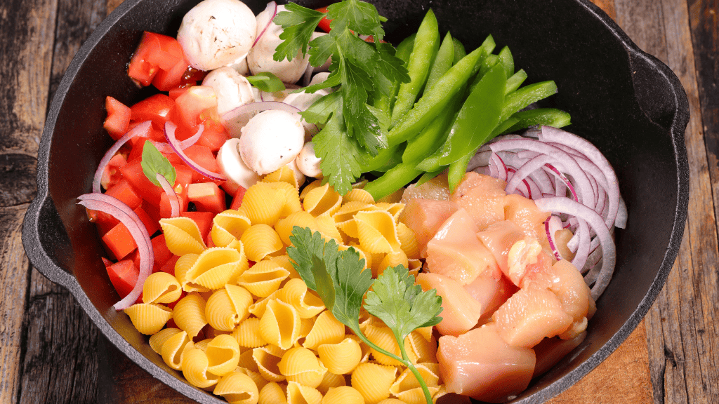 Ingredients for a one-pot meal arranged in a cast-iron skillet, including raw chicken pieces, shell pasta, mushrooms, tomatoes, green bell peppers, red onions, and fresh herbs on a rustic wooden table.