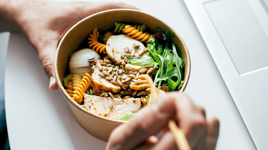 A person holding a bowl containing a balanced lunch of grilled chicken, spiral pasta, boiled egg, arugula, and sunflower seeds, eaten with chopsticks.